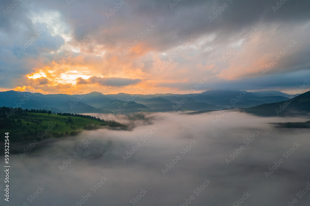 Fog envelops the mountain forest. The rays of the rising sun break through the fog. drone view.