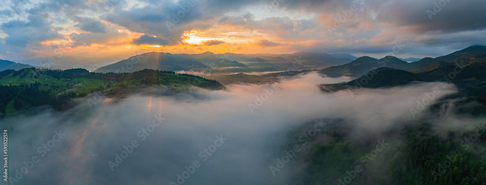 Fototapeta premium Fog envelops the mountain forest. The rays of the rising sun break through the fog. drone view.