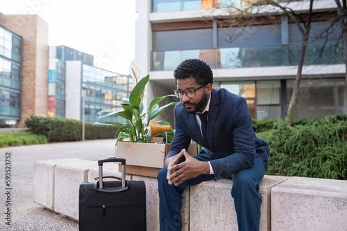 Depressed businessman with suitcase and his belongings sitting outside office building photo