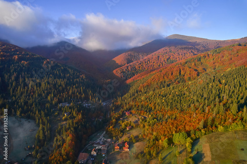Aerial view of mountains at sunrise in autumn in Ukraine. Colorful landscape with mountain road, forest, houses on the hills, sunlight, sky in fall. © Ryzhkov Oleksandr