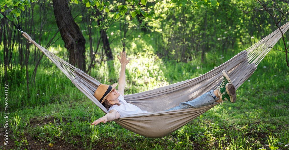 Young happy caucasian woman in a hat lying in a hammock in a green garden enjoying a summer day