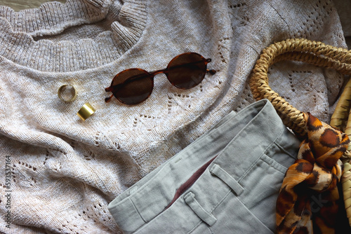 Vintage sweater, neutral trousers, wicker bag, leopard scarf, brown shoes, sunglasses and brass rings on wooden background. Top view.