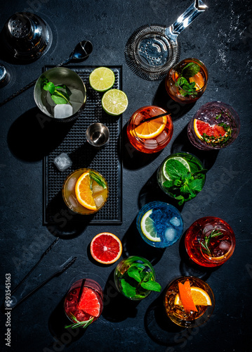 Cocktails set on black bar counter, top view. Mixology concept. Assortment of colorful strong and low alcohol drinks for cocktail party. Dark background, bar tools, hard light, top view