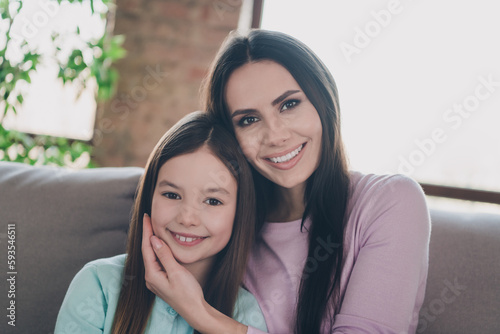 Photo of adorable positive mother hugging her lovely daughter sitting sofa inside room spend time together indoors © deagreez