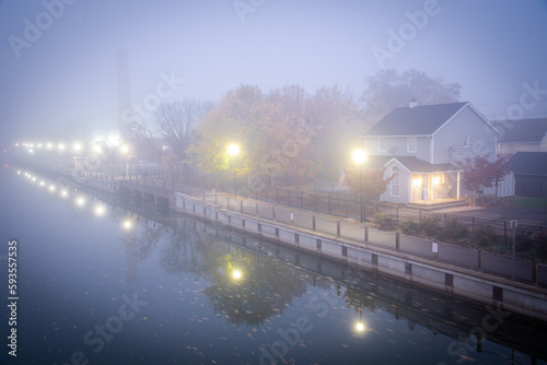 Two story house with American flag in mist foggy early morning along historic Erie Canal with yellow pole lighting, colorful fall foliage in Fairport, Upstate New York, USA