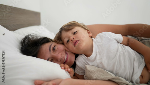 Mother and son laid in bed in embrace looking at camera smiling. Motherhood lifestyle child and mom together