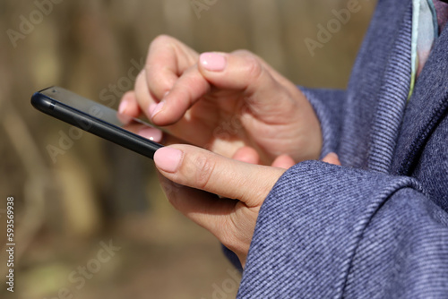 Female hands with smartphone close up on blurred background. Woman in coat using mobile phone stands on a street in spring park