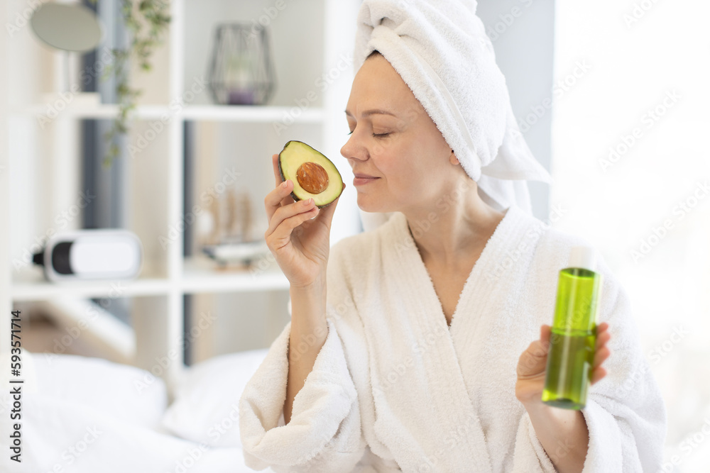 Charming young woman in after-shower outfit enjoying exotic fruit while holding glass vial in bedroom interior. Beautiful female enhancing skin health with avocado facial oil during at-home spa day. Charming young woman in after-shower outfit enjoying exotic fruit while holding glass vial in bedroom interior. Beautiful female enhancing skin health with avocado facial oil during at-home spa day.