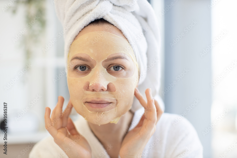 Close up of beautiful calm lady in after-shower wear using cosmetic tissue mask loaded with skincare benefits during at-home spa day. Healthy mature person following weekly facial treatment routine.