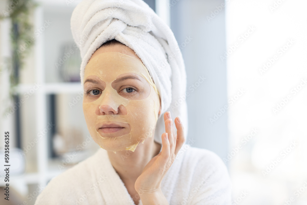 Pretty caucasian female in white robe and towel touching face with applied paper sheet mask while spending free time at home. Young person benefiting beauty care procedures in bedroom interior.