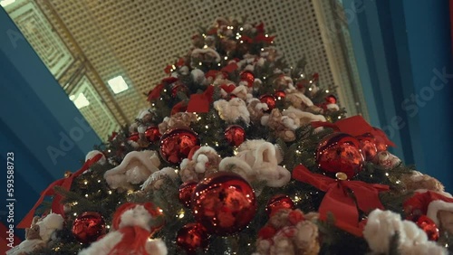 Huge Christmas tree decorated with red bows and balls, plush toys, shining lights. Impressive traditional holiday decorations in a trade center. Winter feast design in a toy store, business center.