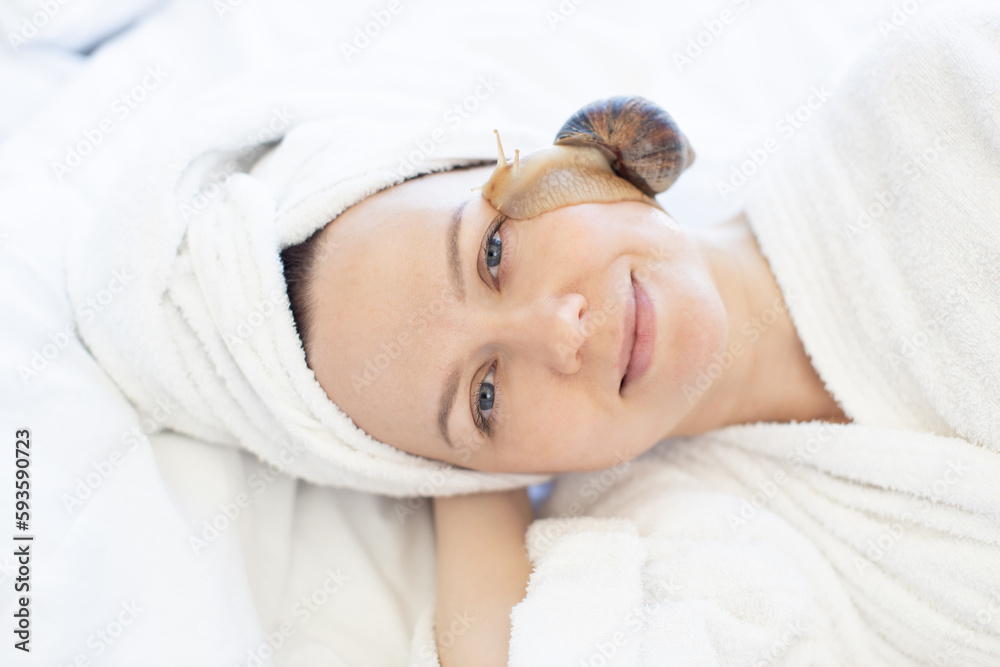 Close up view of young female lying with closed eyes on bed while cute mollusc slithering about on her face. Charming lady in soft towel enjoying snail facial procedure after light massage on bed. Close up view of young female lying with closed eyes on bed while cute mollusc slithering about on her face. Charming lady in soft towel enjoying snail facial procedure after light massage on bed.