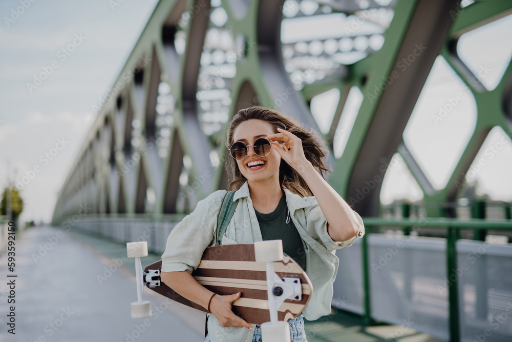 Fototapeta premium Young woman walking on city bridge with skateboard. Youth culture and commuting concept.
