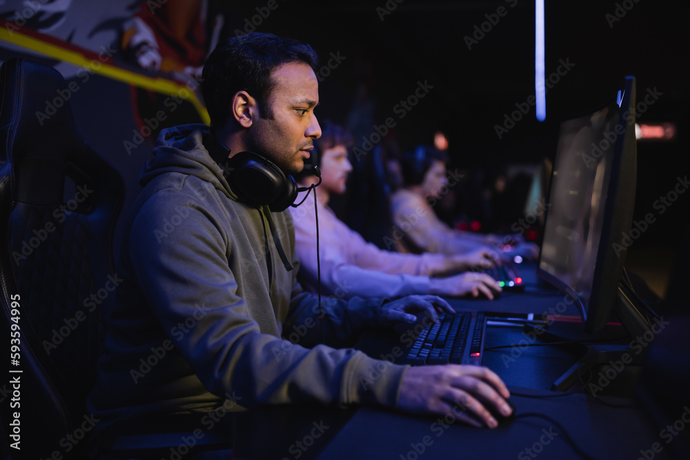 Side view of indian gamer playing video game on computer with blurred team in cyber club.
