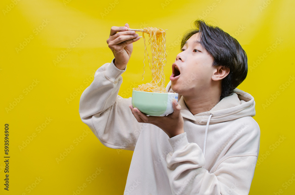 Closeup an Asian man in a beige hoodie eats instant noodles using chopsticks and a bowl, isolated over a yellow background. Closeup an Asian man in a beige hoodie eats instant noodles using chopsticks and a bowl, isolated over a yellow background.