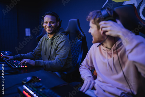 Smiling indian gamer in headphones sitting near blurred friend in cyber club.