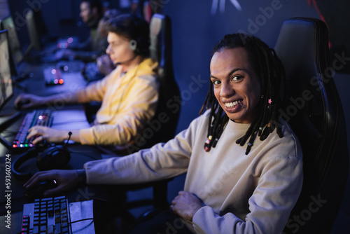 Multiracial gamer smiling at camera near blurred team in cyber club.