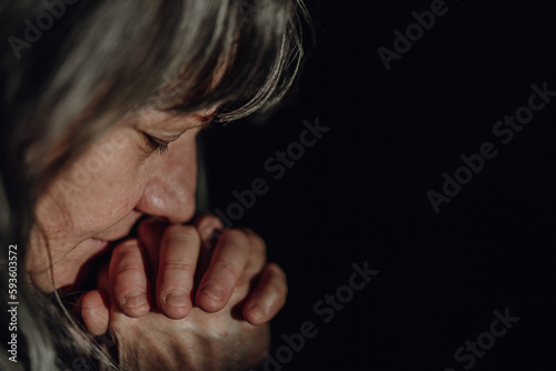 Close-up of unhappy elderly woman praying, black background.