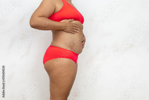 Overweight woman standing half turn massage belly by hands, free copy space, white background. Bare woman in red underwear with cellulite body and stretch marks. Plus size, feminism, self acceptance. © Юля Бурмистрова