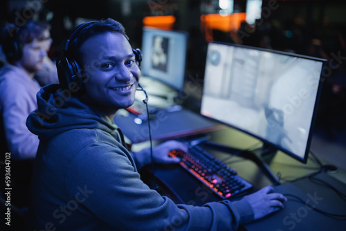 Smiling indian gamer in headphones looking at camera near computer in cyber club with lighting.