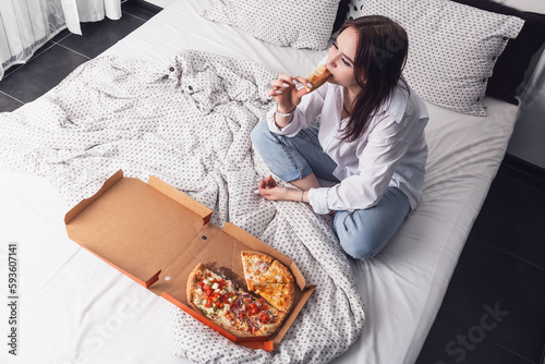 The girl eats pizza while sitting on the bed.