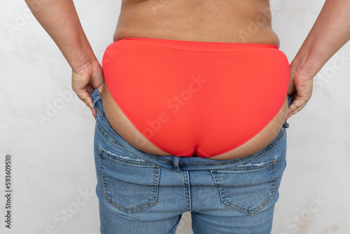 Cropped rearview of fat, obese and overweight female in red lingerie try to pull and get dressed in small size tight jeans, to fit buttocks in clothes on white background. Preparation to summer season