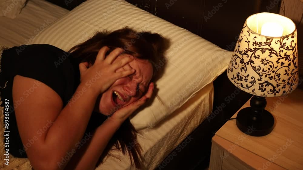Suffering woman lie on pillow in bed and hysterically cry grabbing head and screaming in dark bedroom. Splash out emotions, sob uncontrollably. Emotional breakdown. Night hysterics before sleep.