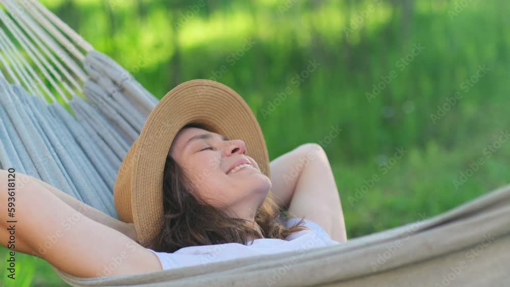 Carefree woman in hat is resting in a hammock in a green garden in sunny summer day. Young girl spending free time, enjoying beautiful weather.