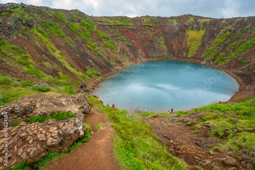 Top view of Kerid Crater Lake in Iceland on a cloudy day with people walking and standing around in the distance. Turquoise waters of Icelandic crater lagoon.