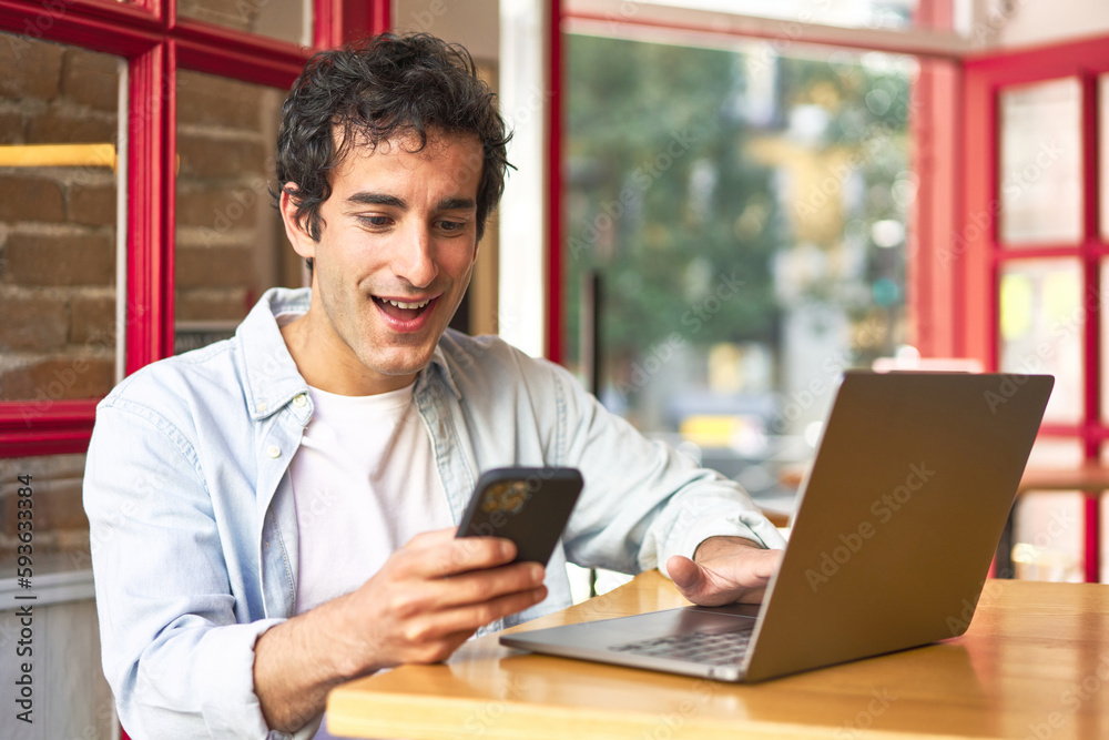 Young man happily multitasking on laptop and phone in cozy cafe. Young man happily multitasking on laptop and phone in cozy cafe.