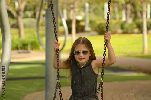 A girl with blond long hair in sunglasses rides on a swing in the spring. Girl 6 years old. Portrait of a child on the playground in the garden