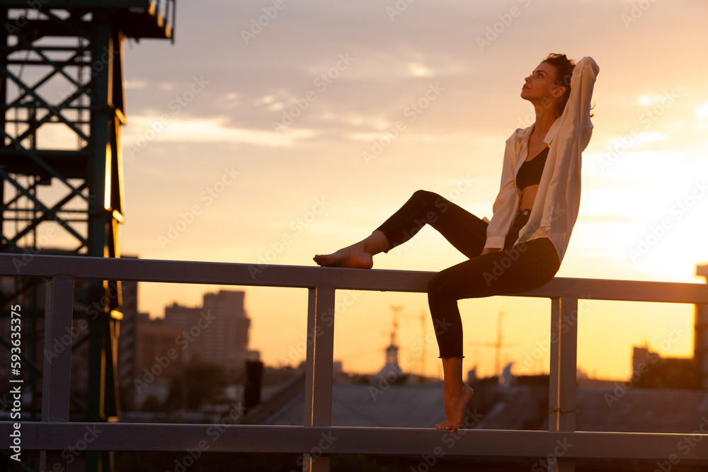 Pensive young brunette sitting on metal railing in rays of setting sun