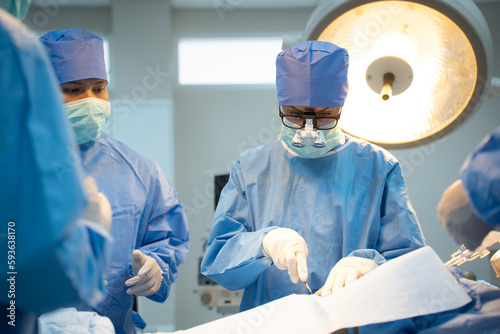 Front view of a Caucasian female surgeon in blue surgical uniform, face mask, and medical loupes, with her team, standing surgery on the patient through a surgical drape in a blurred operating room.