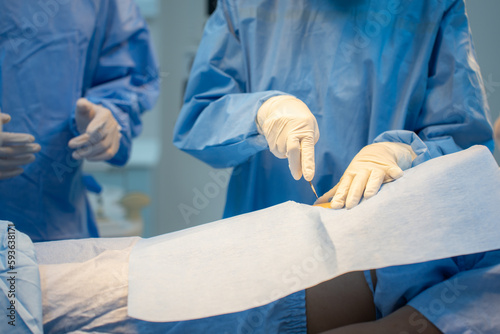 Billede på lærred Selective focus of the hands of surgeons in sterile gloves, wearing blue surgical gown, standing in surgery on the patient through a surgical drape with a blurred team in background in operating room