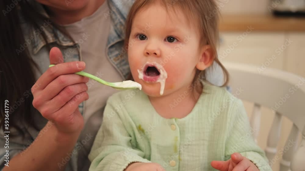 Happy family at home. Mother feeding her baby girl from spoon in kitchen. Little toddler child with messy funny face eats healthy food at home. Young woman mom giving food to kid daughter Happy family at home. Mother feeding her baby girl from spoon in kitchen. Little toddler child with messy funny face eats healthy food at home. Young woman mom giving food to kid daughter
