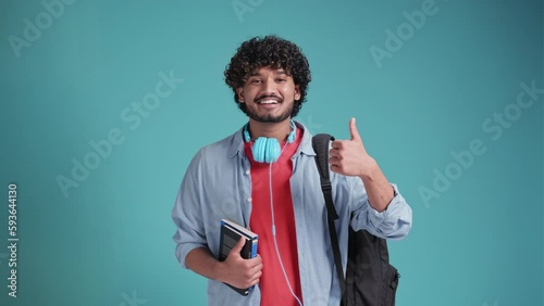Smiling happy young adult arab or indian man student standing isolated on blue background, showing thumbs up like hand gesture giving recommendation advertising good service or result.