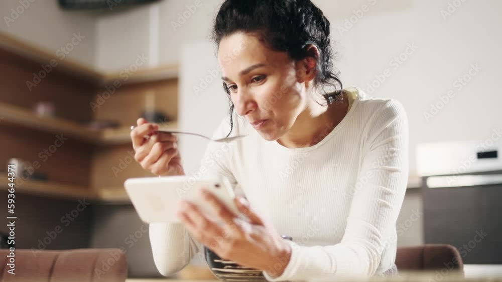 Pretty hispanic woman enjoying beautiful morning with tasty breakfast hold smartphone laughing while watching online funny videos entertainment programs in social media in sunny kitchen at home Pretty hispanic woman enjoying beautiful morning with tasty breakfast hold smartphone laughing while watching online funny videos entertainment programs in social media in sunny kitchen at home