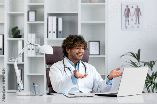 Hindu doctor consulting patients remotely, man working inside modern clinic office, using laptop for online video call, talking and smiling friendly to patient.