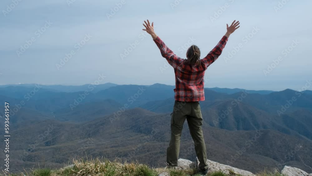Caucasian young man with dreadlocks climbed to top of mountain and spread his arms out to sides from happiness and pleasure. Rear view. Concept of travel tourism and active healthy lifestyle.