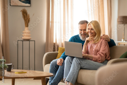 Cheerful Mature Couple Using Laptop Sitting On Sofa At Home