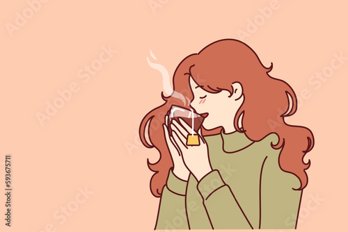 Woman drinks hot tea from transparent glass, enjoying taste of warming drink in cold winter weather. Young girl in sweater feels cozy and drinks tea drink take break to rest from work 