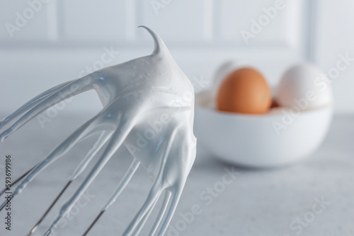 Whisked egg whites - whipped Italian meringue on a wire whisk and eggs on a gray background. copy space.