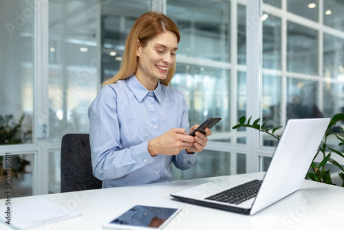 Successful mature businesswoman boss using phone at workplace inside modern bright office, office worker typing online message on smartphone smiling