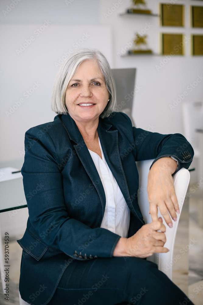 Smiling mature female entrepreneur with gray hair in formal suit sitting on chair and looking at camera in light room