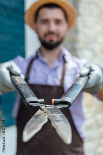 Stylish man-gardener with garden scissors in hand standing in front of garden house