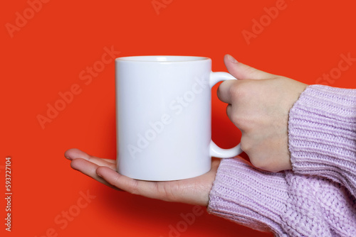 A woman's hand holds a white cup with a hot drink on an orange background. The hand of a woman dressed in a pink sweater holds a ceramic cup on an orange background