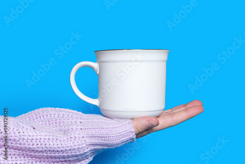 A hand holds a white cup with a hot drink on a blue background. The hand of a woman dressed in a sweater holds a large ceramic cup on a bright blue background. The concept of drinking hot drinks