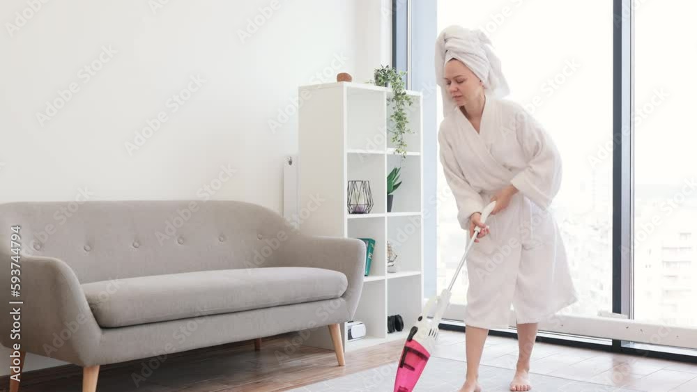 Smiling barefoot lady after shower using lightweight stick vacuum for daily clean-up of airy studio room. Joyful female homeowner saving time and energy while hoovering with easy handheld device. Smiling barefoot lady after shower using lightweight stick vacuum for daily clean-up of airy studio room. Joyful female homeowner saving time and energy while hoovering with easy handheld device.