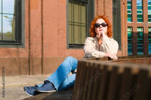 Attractive red-haired woman with sunglasses sits on a bench in a business district. Woman on a spring walk. Confident middle-aged woman. © Galina