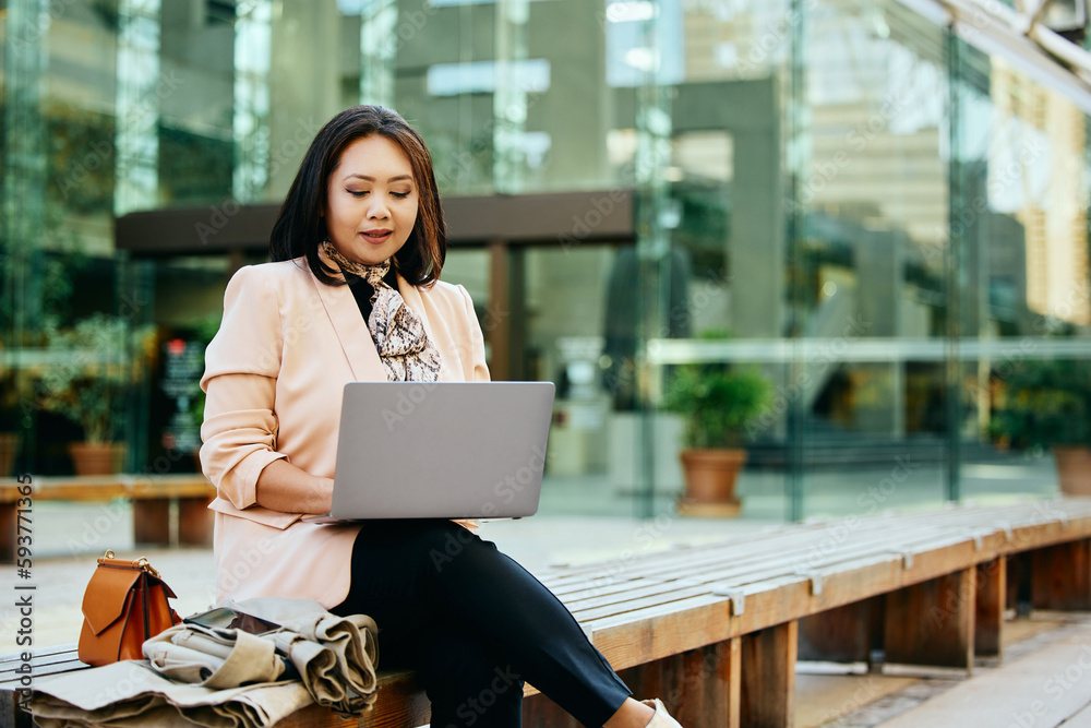 Fototapeta premium Asian businesswoman surfing the net on laptop while sitting on bench in city.